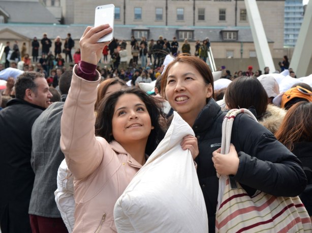 Two women are standing together as they take a selfie at Nathan Phillips Square during the pillow fight that was held there. One woman is holding a phone, the other woman is holding a pillow. 
