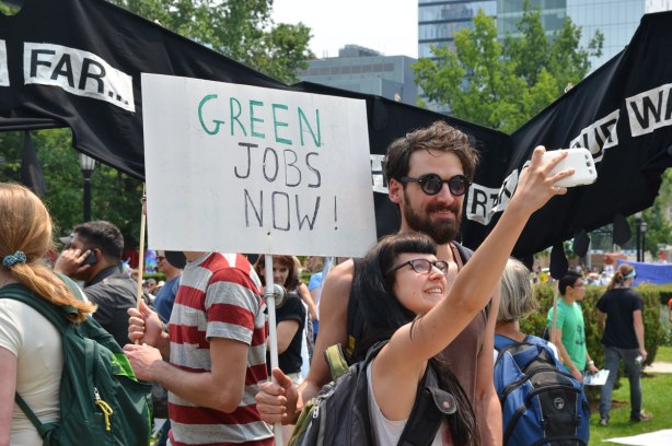 A couple is taking a selfie at a protest march. SOmeone is holding a sign that says Green Jobs Now behind them. 