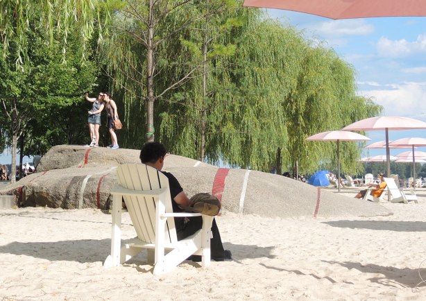 A man is sitting on a white chair under a pink umbrella on Sugar Beach. Behind him are two people standing on a rock taking a selfie together 