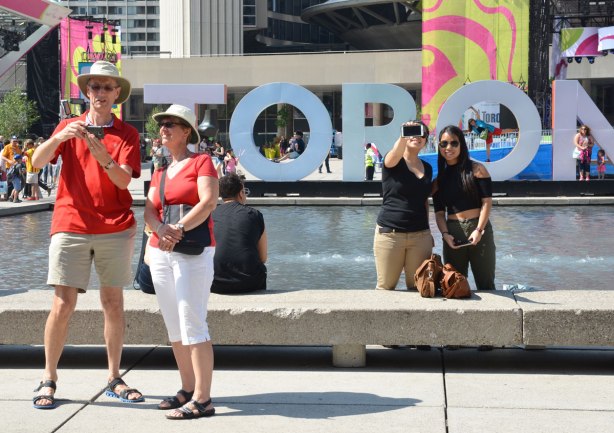 A couple dressed in red and white are taking a selfie beside each other in front of the 3D Toronto sign at Nathan Phillips square. Two other people are to the right, also taking pictures. They could be taking a selfie or their camera could be pointed away from them. 