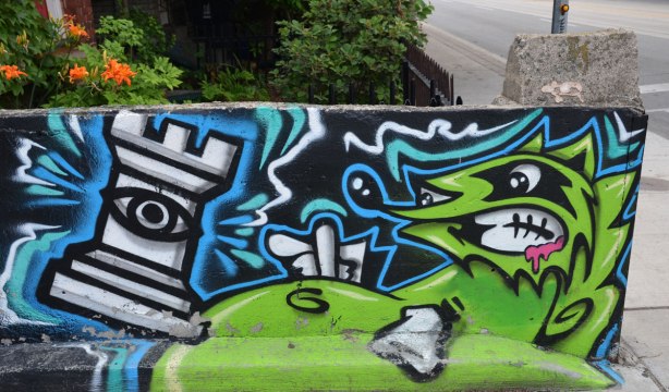 a low concrete fence is painted with graffiti street art, a green snarling green raccoon creature is acting out against a chess piece, a castle or rook