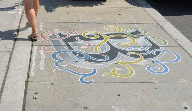 The letter B is painted on a sidewalk in black, with blue, gree, yellow and swirls like ribbons wrapped loosely around the letter