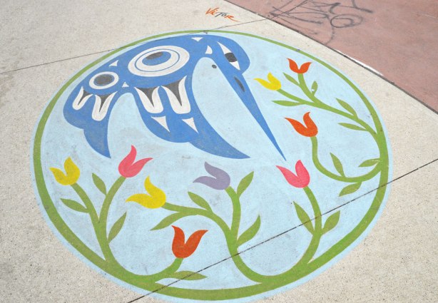 A painting on a sidewalk of a hummingbird done in Inuit style in blue and white, also some flowers, all enclosed in a green circle. 