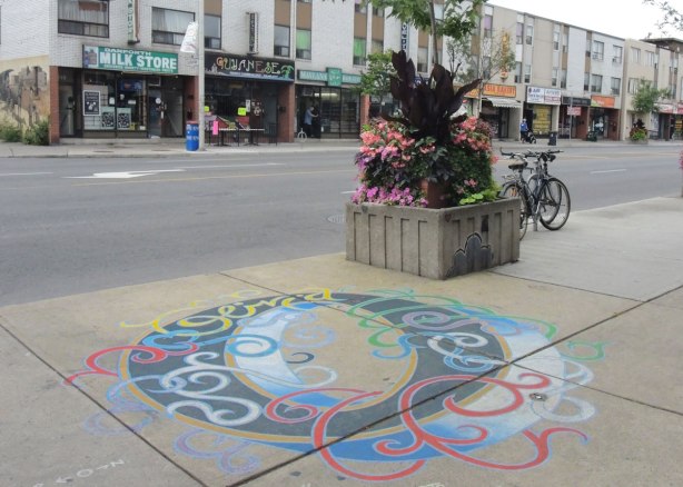 The letter O is painted on a sidewalk in black, with blue, green, yellow and swirls like ribbons wrapped loosely around the letter