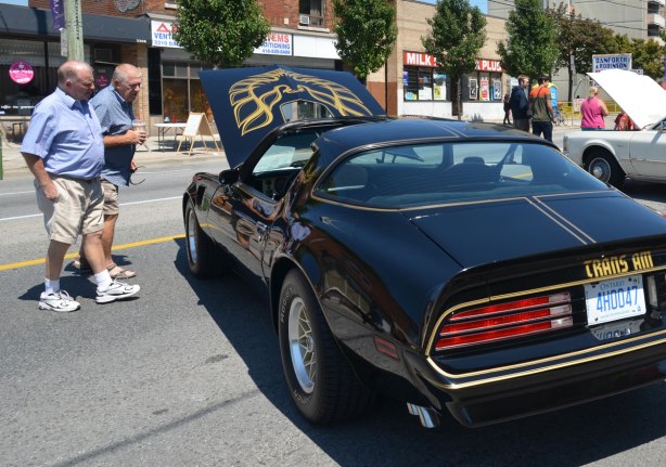 picture taken at a car show - two men are talking together and looking under the hood of a black Trans Am