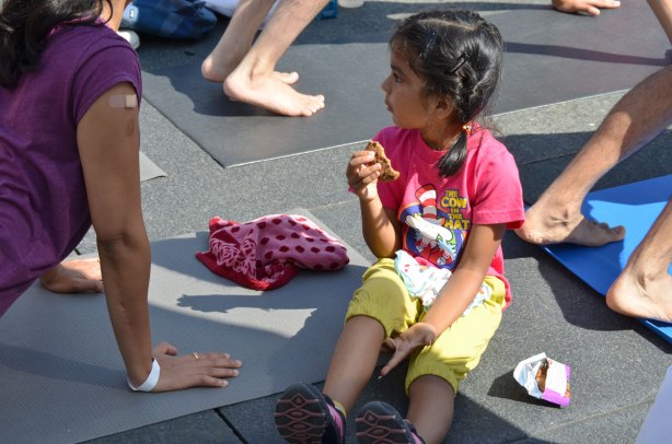 men and women at a yogathon - doing yoga outside in a large group, young girl sitting on the ground and eating a cookie while her mother does yoga. 