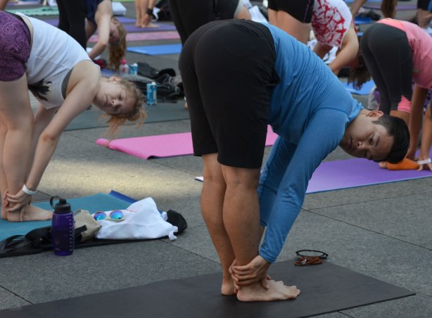 men and women at a yogathon - doing yoga outside in a large group - a man stands with his hands around his ankles, knees straight. 