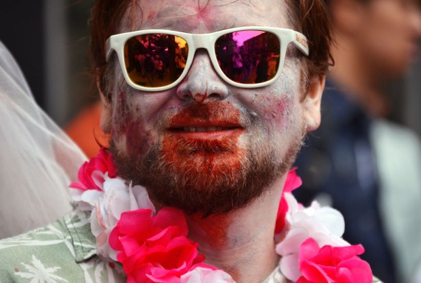 people dressed up as zombies - a man with a beard, bloody face, and white framed sunglasses. He is wearing a pink and white garland of fake flowers around his neck