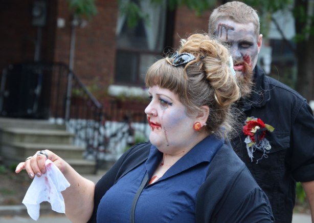 people dressed up as zombies - two zombies, a woman in a blue dress waving a white hanky and behind her a man looking directly at the camera