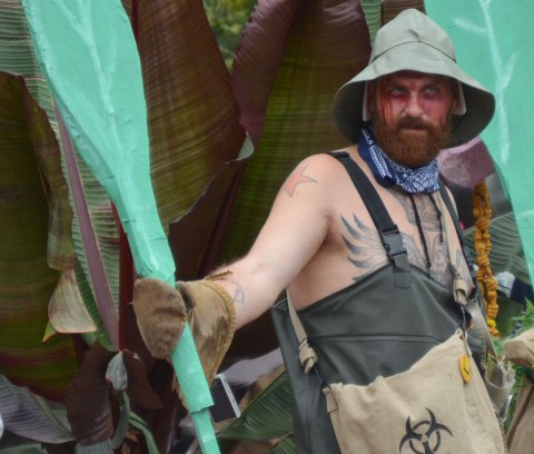 people dressed up as zombies - man with green hat and holding two large leaf like poles, standing in front of a leafy plant