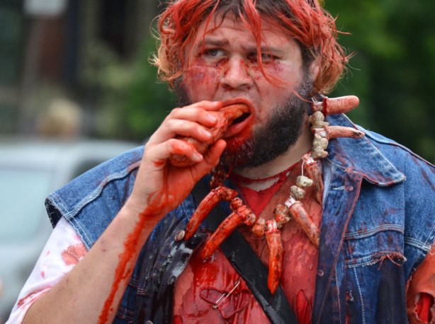 people dressed up as zombies - a man with a necklace made of fingers and bones, covered in blood as he munches on a finger. 