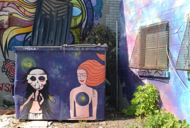 two women painted on a purple dumpster. The one on the left is by mska and the woman has a skull mask on. The other is by paula prezende and is a woman with long red hair but with a big hole in her chest.