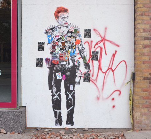 A partial painting of a red head man, life size or close to it, on a white wall that people have added stickers to. The stickers cover all of his torso 