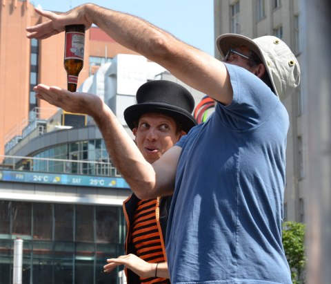 A busker, Funnykito, has a volunteer on stage, The man (volunteer) is holding a beer bottle upside down between the palms of his two hands