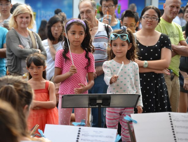 Two young girls take a turn at conducting a small group of musicians playing string instruments at an outdoor festival 
