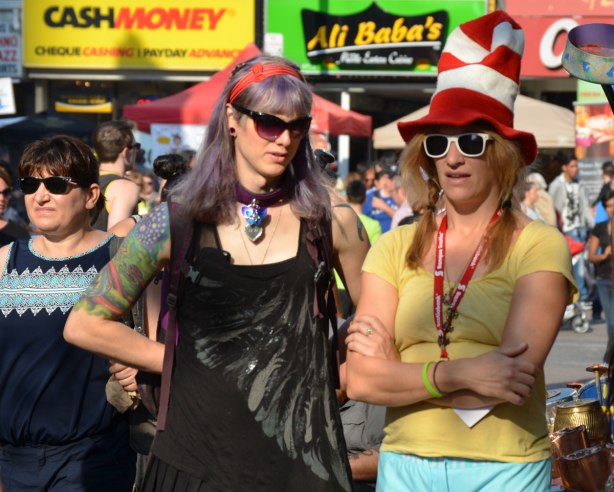 Two people watching a performance at a street festival. One of them is wearing a large red and white Dr. Suess hat. 