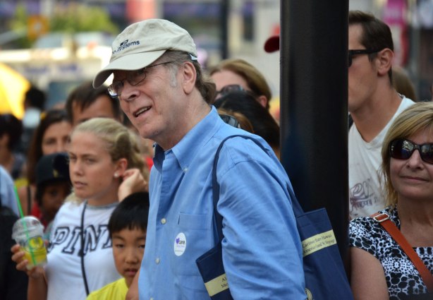 A man in a blue shirt and beige baseball cap watches in fascination at a performance at a street festival. There are other people in the crowd around him. 