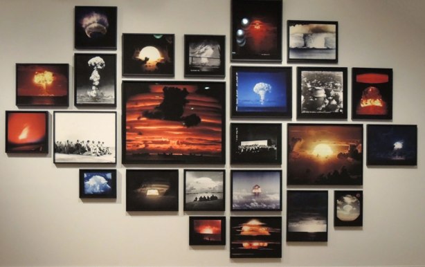 A collage of photos of nuclear blast mushroom clouds. There are about 25 colour and black and white photos hanging on a wall of the Art Gallery of Ontario