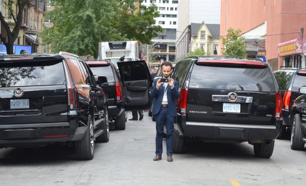 A short man is taking pictures of a crowd (directly towards the camera) or else he is taking a selfie with a lot of black Cadillac Escalades behind him. 