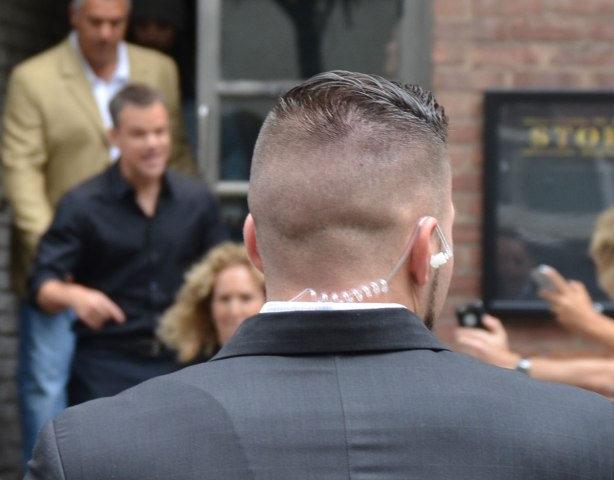 The back of the head of a security guy. His head is shaved except for the top part. He is wearing a wire. Matt Damon and another security guy are out of focus in the background. 