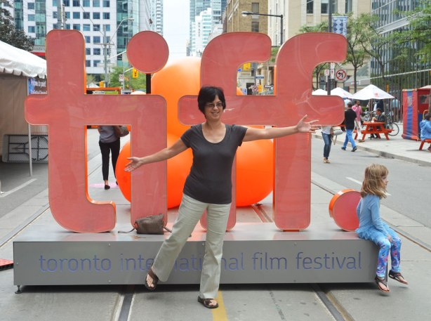 A middle aged Asian woman poses in front of the orange tiff 3D sign on King Street. A girl is sitting on the corner of it, looking off the picture to the right. 