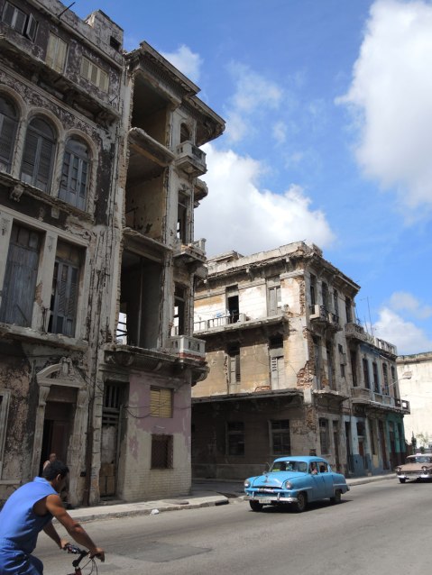 picture taken inthe old part of the city of Havana Cuba - old colonial style three storey buildings that are empty and abandoned.  Two old American cars are driving past them. 