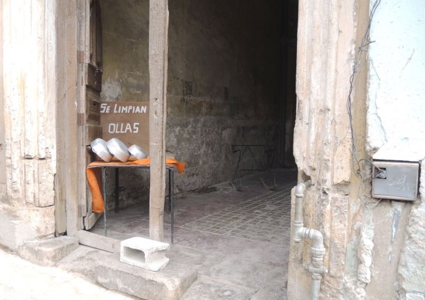 picture taken in the old part of the city of Havana Cuba - A doorway of a rather empty store.  Three bowls are on an orange cloth on a table.  The sign by the bowls says se limpian ollas (we wash bowls). 