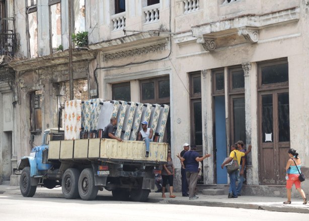 picture taken inthe old part of the city of Havana Cuba - delivering mattresses.  A truck with seven mattresses is parked in front a building.  Men are talking on the sidewalk. 