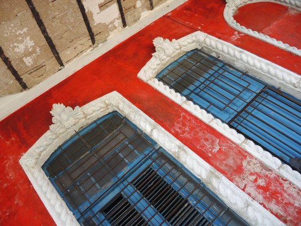 picture taken inthe old part of the city of Havana Cuba -  photo taken on an angle and looking up at the tops of two windows with ornate white window frames against a bright red wall. 
