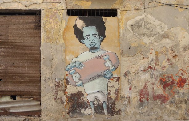 picture taken inthe old part of the city of Havana Cuba - graffiti on a wall.  A man with curly black hair and a blue face is holding a skateboard. 