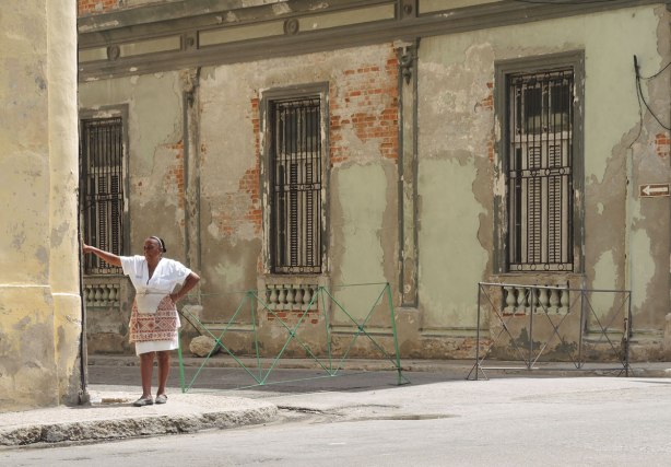 picture taken inthe old part of the city of Havana Cuba - a woman in a white blouse and white skirt and multicoloured apron leans agains the corner of the exterior of a building.  A weathered old building is behind her. 