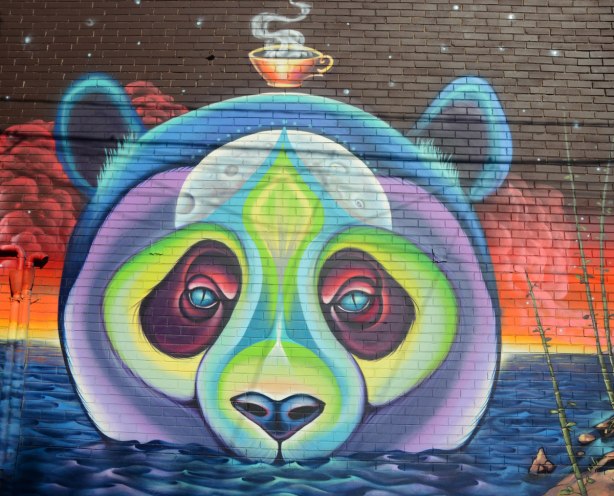 The face of a bear, perhaps panda bear, but in purples and greens. It is in water up to its nose. A steaming tea cup floats above its head. 