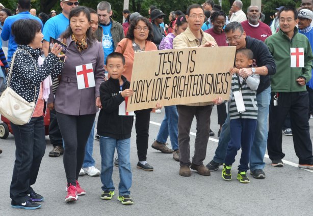 Two boys are holding a brown banner that says Jesus is holy, love, rightousness. A group of Asian people are walking with them in a parade. 