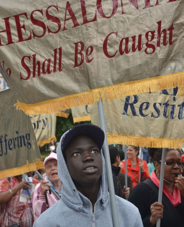 A young black man is holding a banner in a parade. He is looking upwards. Other people around him are also holding banners. The banners are gold and each one has the name of a book of the bible on it.