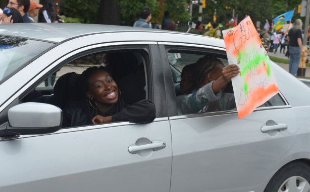Two women in a car. The driver is making a smiley face. The women in the backseat is holding a sign out the window that says 