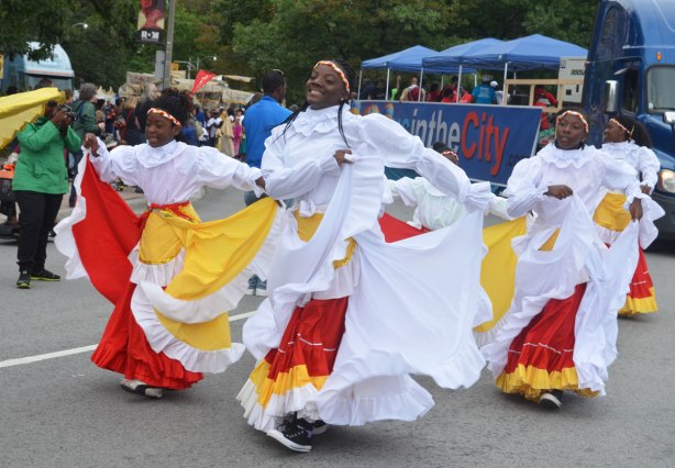 Four women wearing long ruffled skirts in white, red and yellow, swishing their skirts side to side as they walk in a parade 