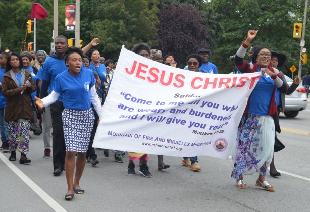 Two women in blue t-shirts are holding a banner in a parade. Others are walking with them. The banner says Jesus Christ in large red letters. There are smaller words in blue below that are a verse from Matthew that starts with Come to me all you who are weary and burdened and I will give you rest. The group is from the Mountain of Fire and Miracles Ministry
