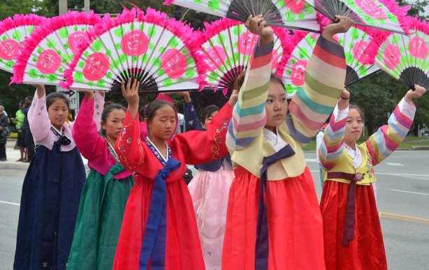 A line of Japanese women in kimonos are carrying large open fans above the heads. The fans have big pink flowers on them as well as a pink feathery border. 