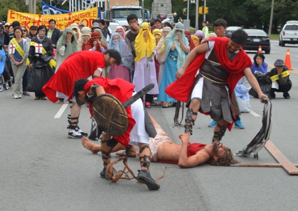 Four men are reenacting Jesus being taken by the Romans for crucifixion. A man is on the ground while the Roman soldiers whip and beat him. A cross is lying on the ground too. A group of upset women onlookers (part of the acting) follow behind. Part of the Jesus in the city parade. 