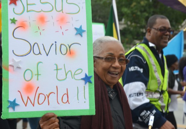 An older black woman with a very big smile is holding a sign that says Jesus is the Savior of the world. She is walking in the Jesus in the City parade in Toronto.