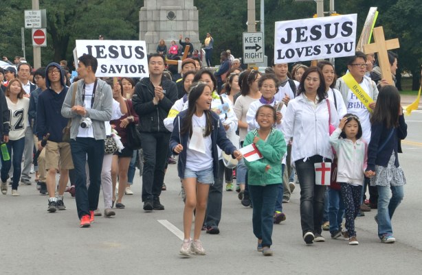 A group of people, mostly young and mostly Asian, are walking in a parade. One is holding a sign that says Jesus loves you. Another person is holding a sign that says Jesus is Savior. 
