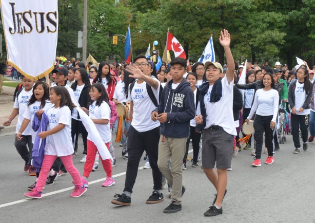 A group of young people are walking in a parade. Some are waving to the camera. One girl is holding a banner with just the word Jesus on it. 