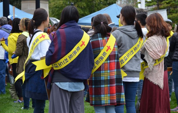 That backs of 4 young women. THey are all wearing sweaters and yellow sashes. The sashes have Korean or maybe Chinese lettering on them as well as a red cross. 