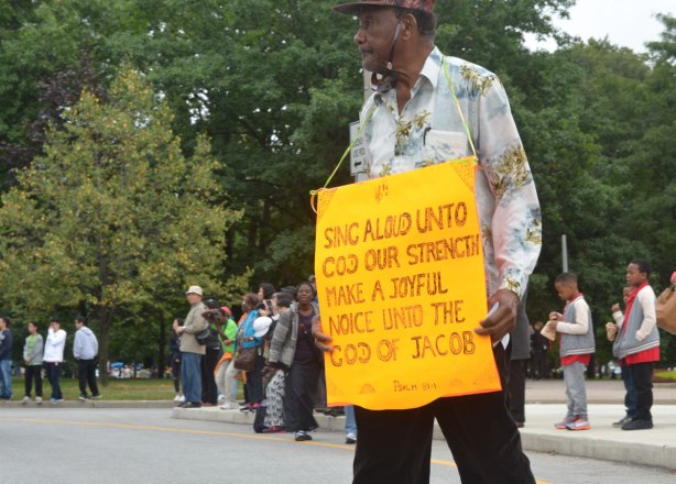 A black man is wearing a yellow sign around his neck. It says Sing aloud unto God our strength make a joyful noise unto the God of Jacob. 