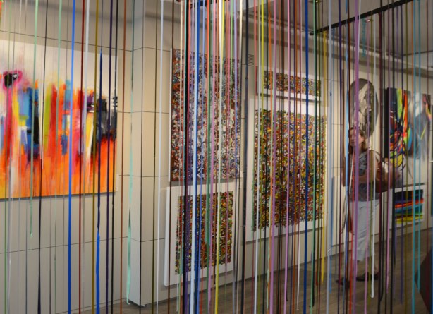 Curtains of long strips of dried acrylic paint in many bright colours hang from the ceiling of an art gallery. 