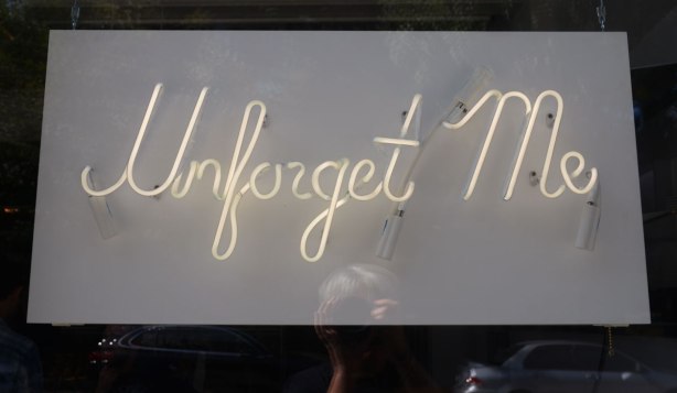 In sursive writing the words Unforget Me in neon tubing to make a sign that is hanging in a store window. 