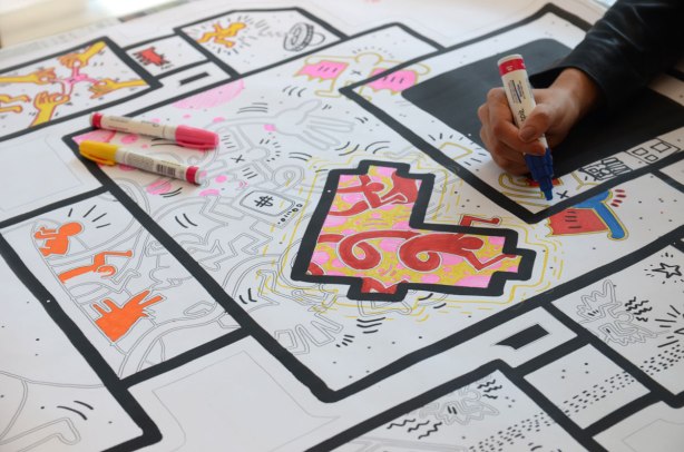 A large lovebot on paper is on a table. Black lines on white paper. It is more than a meter high. There are shapes in the middle and it is designed to be coloured. A hand holding a black sharpie is also in the picture, colouring part of the lovebot 