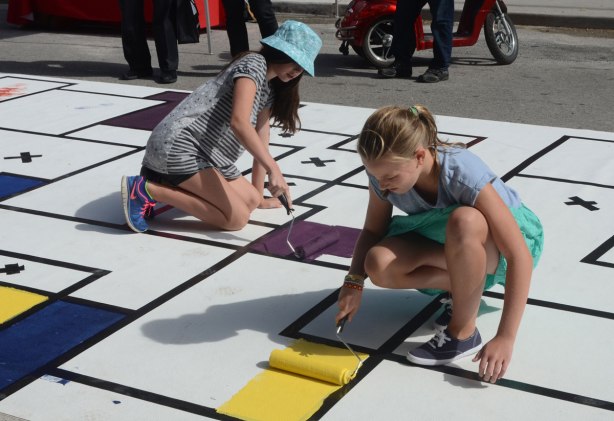 painting a large Mondrian-like painting on the street. A large mat is laid out along Frederick Street and students have marked off squares and rectangles with tape. People are painting the shapes in red, orange, yellow, green and purple. 