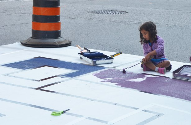 painting a large Mondrian-like painting on the street. A large mat is laid out along Frederick Street and students have marked off squares and rectangles with tape. People are painting the shapes in red, orange, yellow, green and purple. A young girls is using a small roller to paint purple in this picture