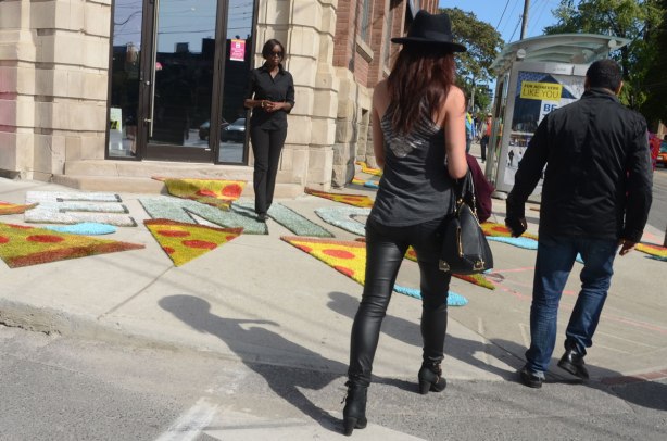 A man and a woman are crossing the street. They are close to the sidewalk on the other side of the street. On that sidewalk are a number of carpets that look like wedge shaped pepperoni pizza slices. Each carpet is just over a meter long. They are in front of a shop that sells appliances. 